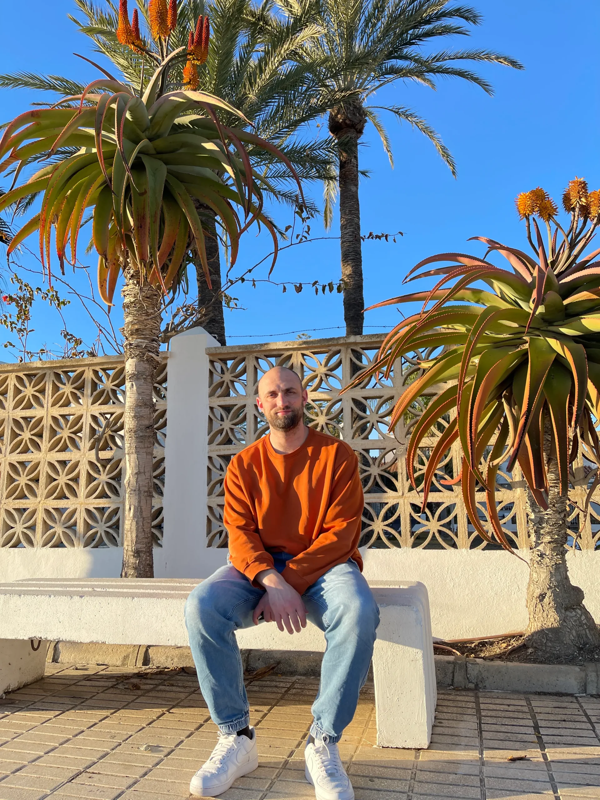A picture of Christopher Cardoen sitting on a bench in between palm trees.